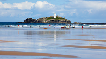 Godrevy Surfing This landscape photograph shows Godrevy Beach in Cornwall, England, during a summer morning. The main subject of the image is Godrevy surfing at Gwithian, with several surfers and beachgoers visible along the shallow shoreline where the wet sand creates clear reflections of the people and flags. In the background, the sea meets Godrevy Island, which features the prominent Godrevy Lighthouse, a well-known landmark of the Cornwall coastline in the United Kingdom. The calm summer light and blue skies enhance the vivid colours typical of the region, while the gentle waves underline the beach's popularity as a surfing destination.